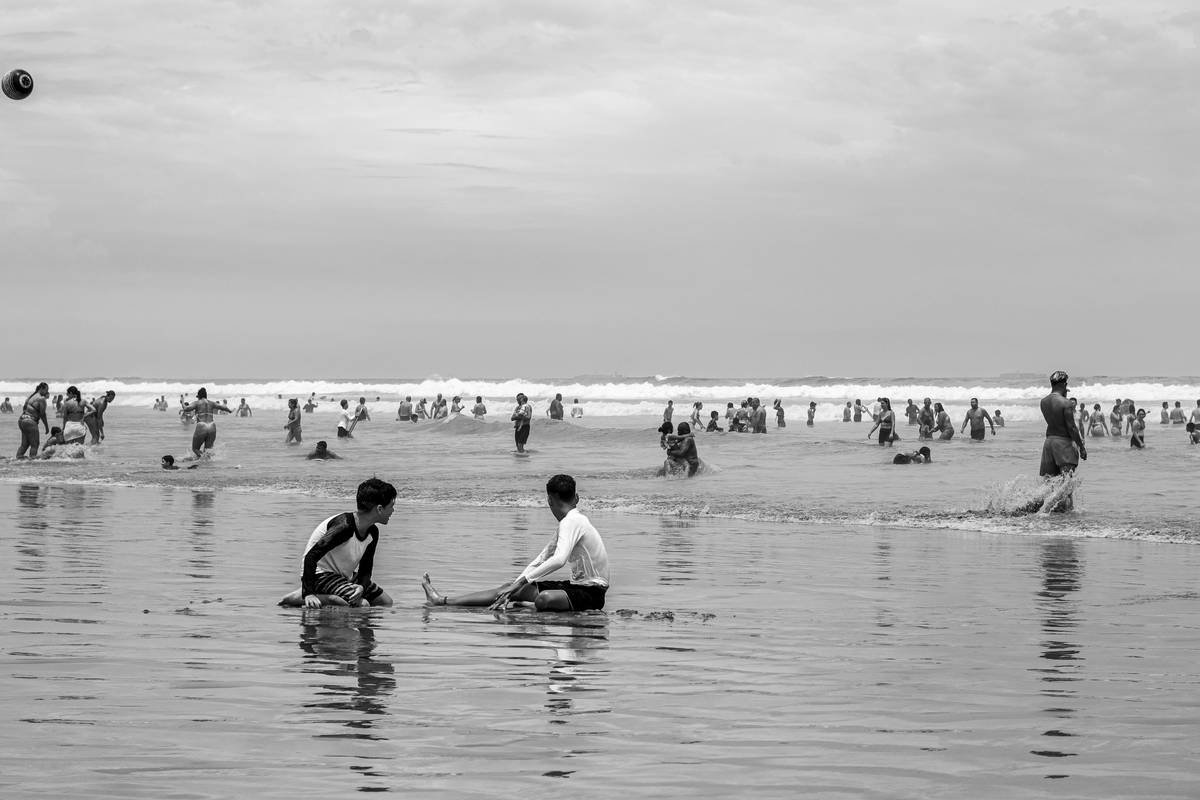 A group of people are enjoying a day at the beach, with many swimming and playing in the ocean waves. In the foreground, two young boys are sitting in the shallow water, looking at each other.