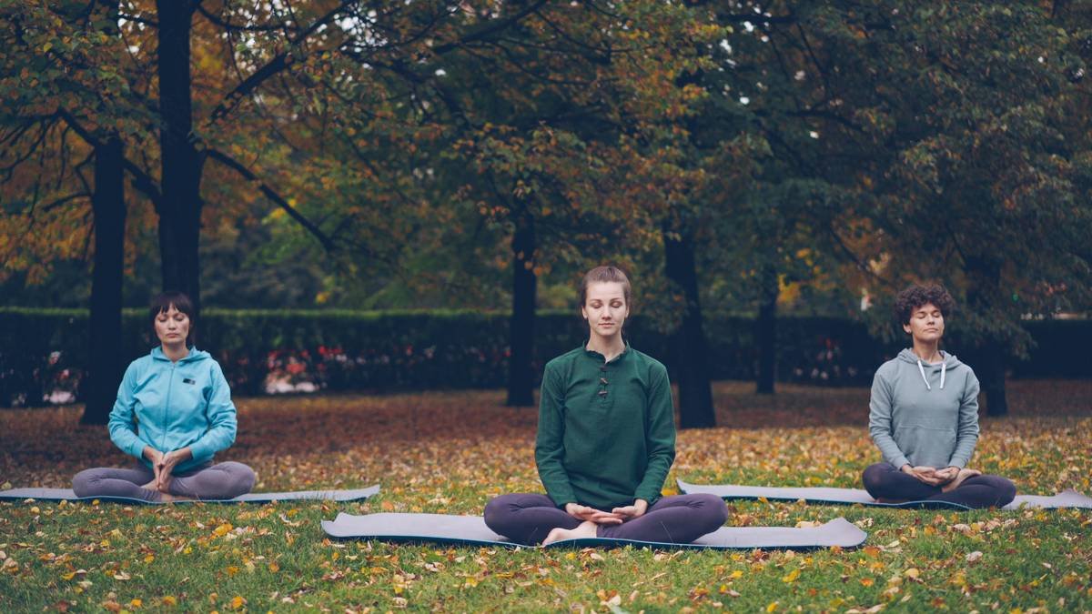 Female yoga instructor is showing stretching exercises then relaxing in lotus position with closed eyes sitting on mats during open-air practice in city park.
